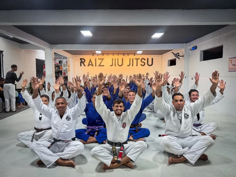 Turma do CT Raiz Jiu-Jitsu 46 sentada no tatame com as mãos levantadas